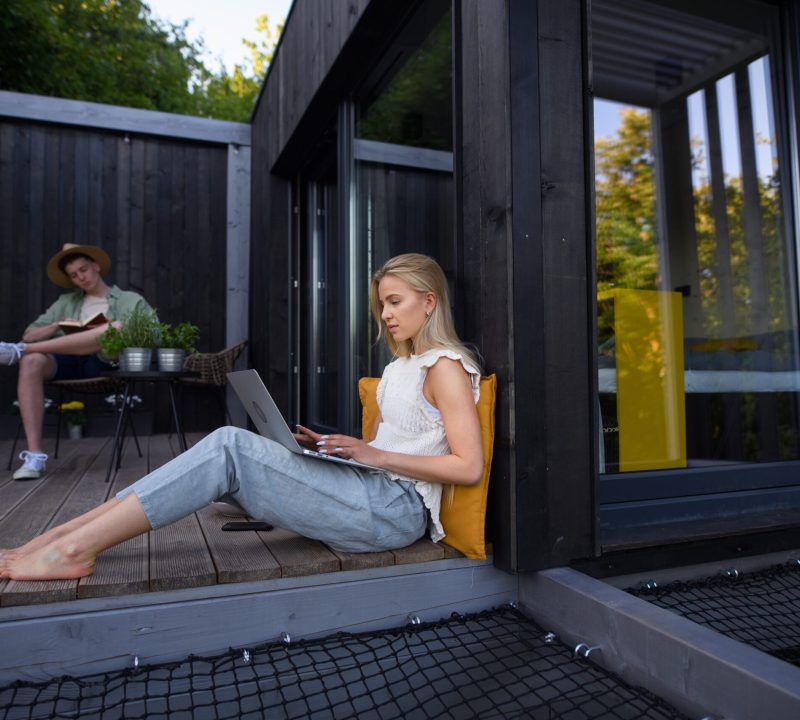 A happy young couple with laptop resting outdoors in a tiny house, weekend away and remote office concept.